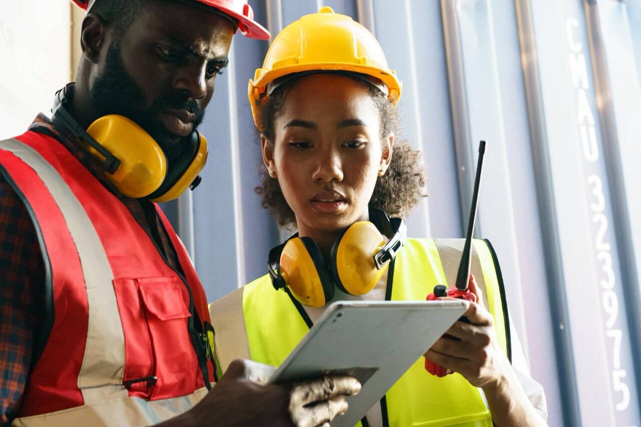Two industrial workers in PPE reviewing site information on a tablet at a logistics facility, representing corporate and industrial operations